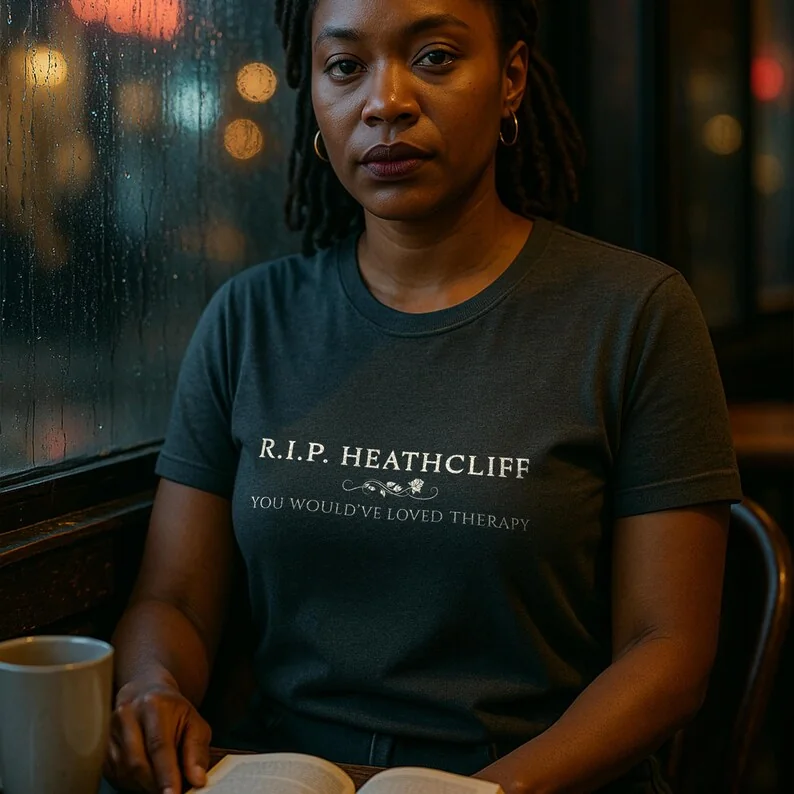 A woman with dark, curly hair sits by a rain-streaked café window wearing a Pepper Comfort Colors t-shirt. Her expression is pensive, with a vintage novel open in front of her. The moody café lighting and raindrops on glass create a gothic, introspective vibe.