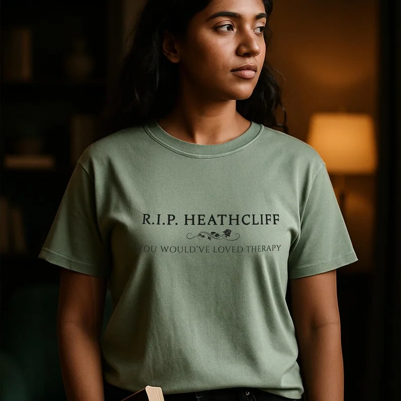 A young woman stands beside a stack of old books. She wears a Bay-colored Comfort Colors shirt . The lighting is warm and melancholic, her gaze distant. The scene suggests quiet reflection, perfectly matching the emotional core of Wuthering Heights.