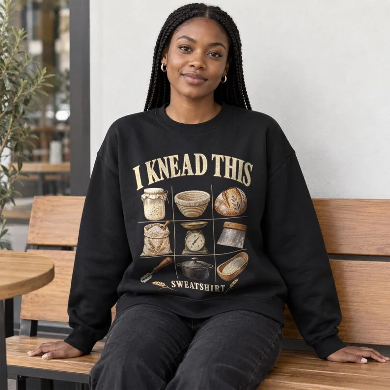 Woman wearing a black crewneck sweatshirt featuring the I Knead This sourdough baker design with nine vintage baking illustrations.