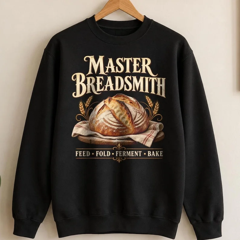 A black long-sleeve sweatshirt featuring a graphic of a round loaf of bread on a cloth-lined wooden board. The text reads MASTER BREADSMITH above the bread and FEED • FOLD • FERMENT • BAKE below the bread in a gold-toned font.