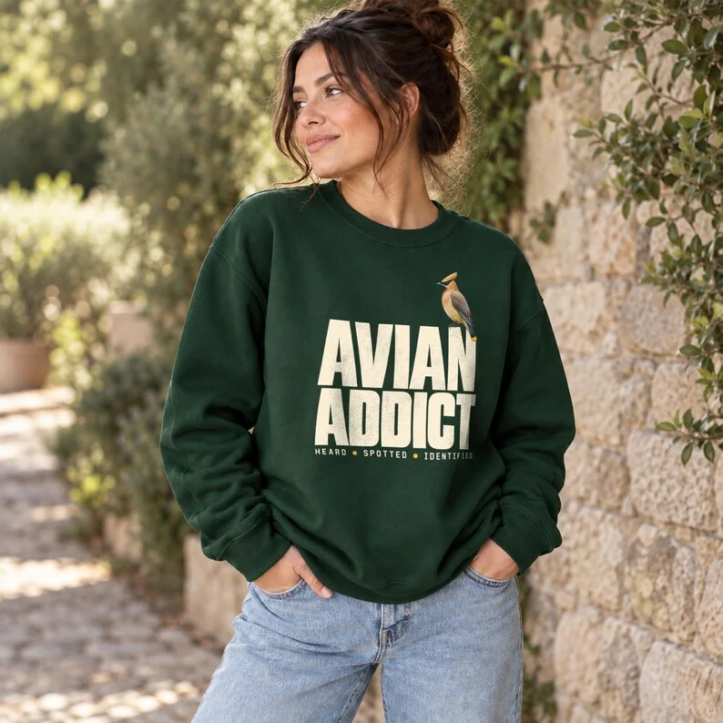 Forest green Avian Addict sweatshirt on woman outdoors, with cedar waxwing birding design and bold typography.