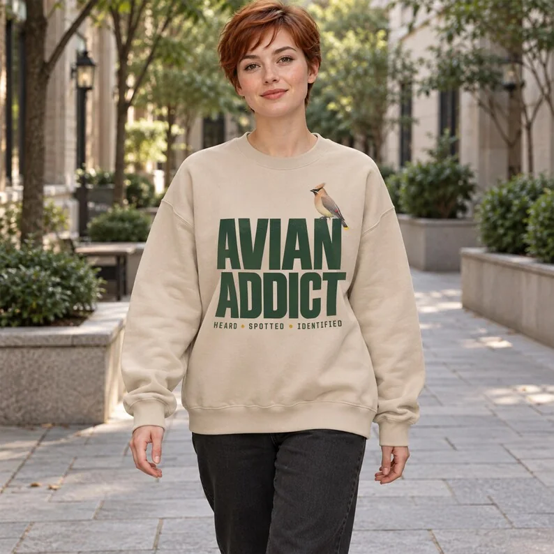 Sand Avian Addict birdwatching sweatshirt on woman outdoors, featuring cedar waxwing and Heard Spotted Identified text.